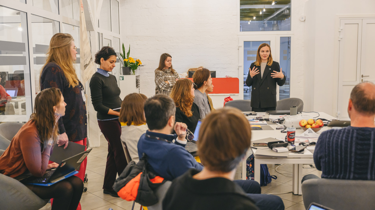 Sieviete melnā žaketē ar žestiem uzstājas gaišā telpā, kamēr grupa cilvēku klausās, daži sēž pie galda ar klēpjdatoriem un āboliem, citi stāv.