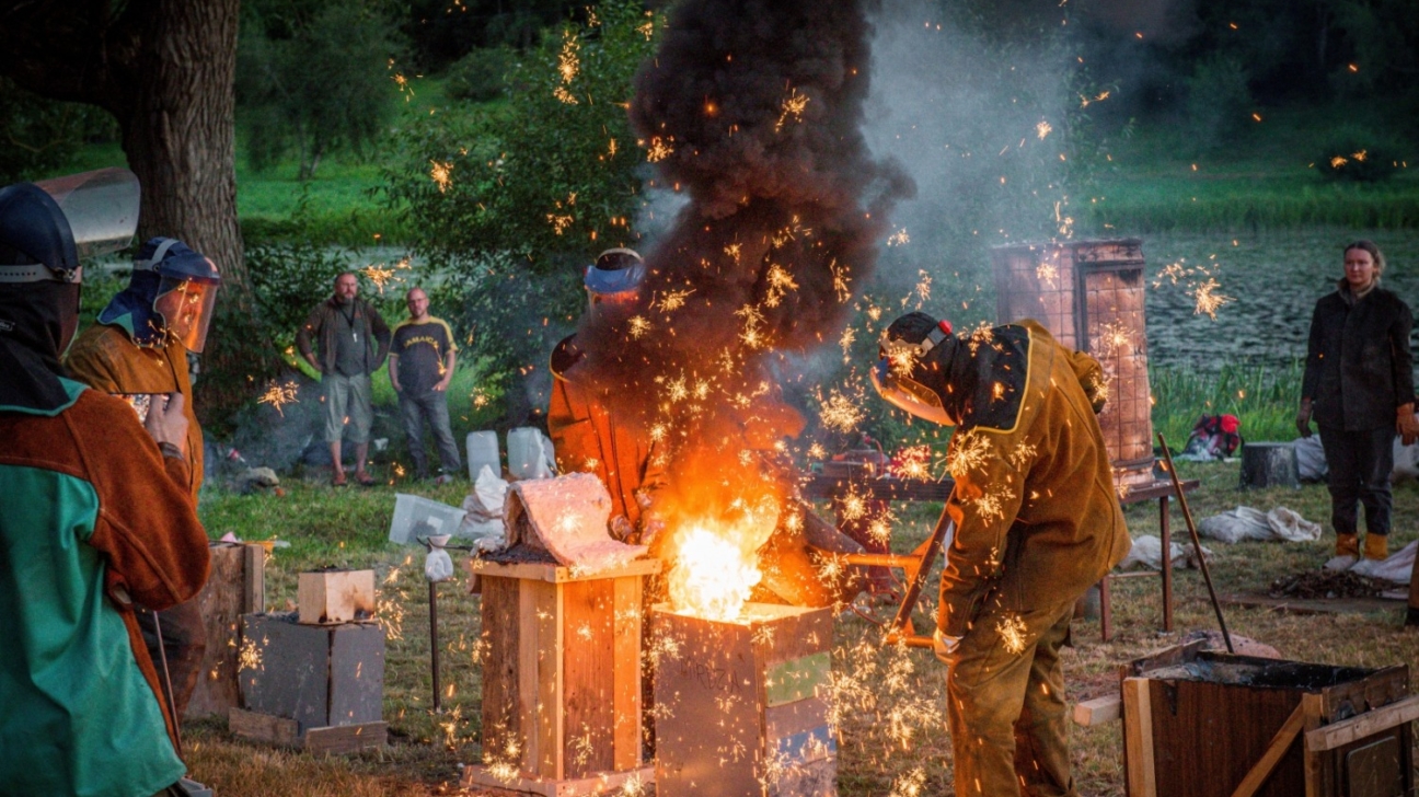 Cilvēki aizsargtērpos veic metāla liešanu ārā, radot spilgtas dzirksteles un melnus dūmus. Viņi stāv pie kvēlojošas krāsns, fonā pie ūdenstilpes vēro citi cilvēki.