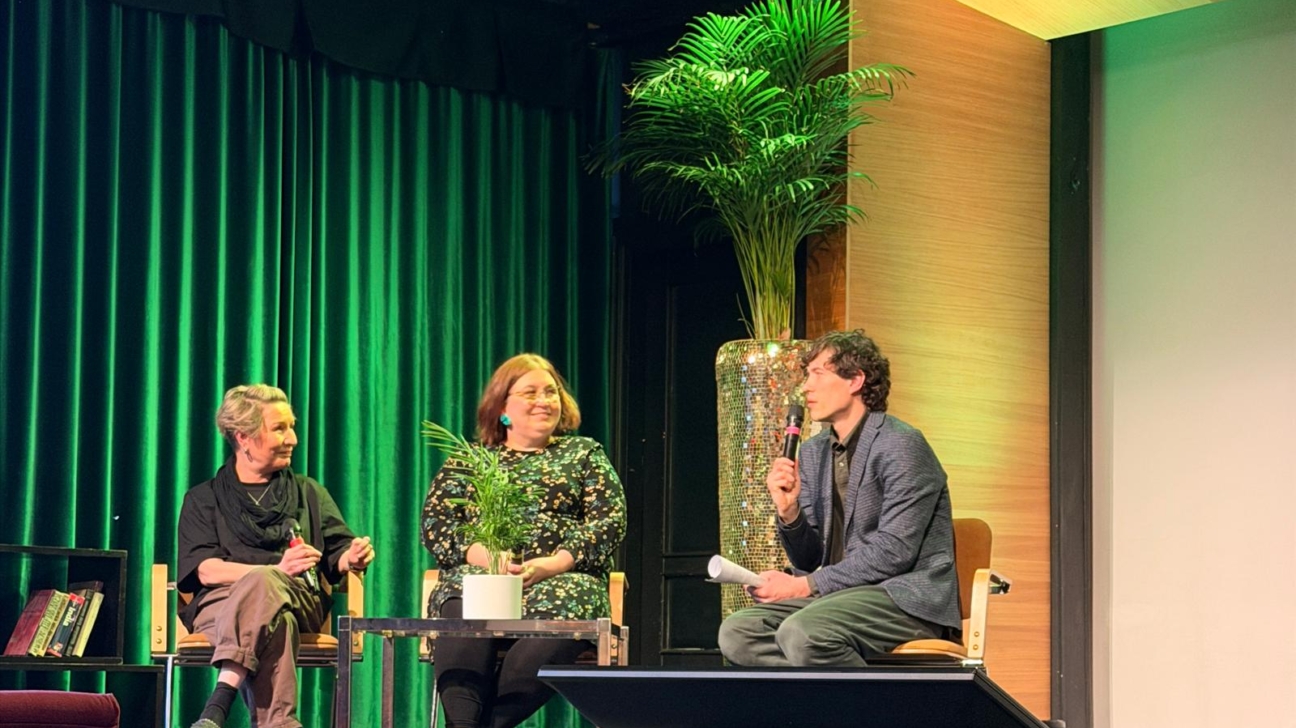 Trīs cilvēki uz skatuves: divas sievietes sēž un vīrietis ar mikrofonu rokās runā. Aizmugurē ir zaļi aizkari un liela palma.