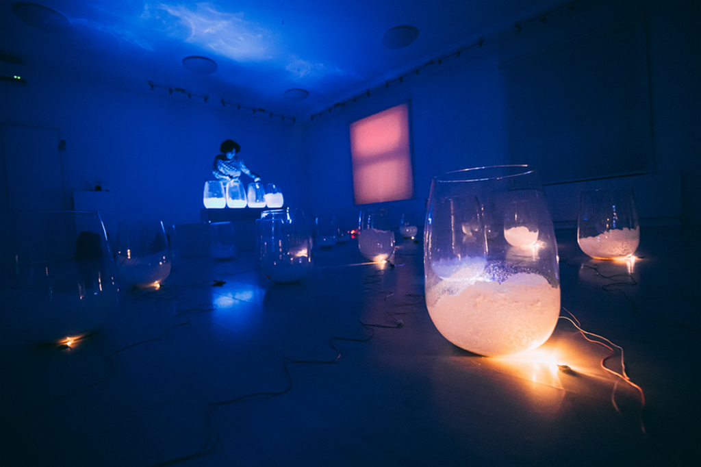 Tumsā, zilganā gaismā izgaismotā telpā uz grīdas ir izvietotas daudzas stikla glāzes ar baltu pildījumu, katra apgaismota ar siltu gaismiņu. Aizmugurē, uz paaugstinājuma, sēž cilvēks un strādā pie vairākām spīdošām stikla tvertnēm, kamēr uz sienas redzams sarkans gaismas laukums.