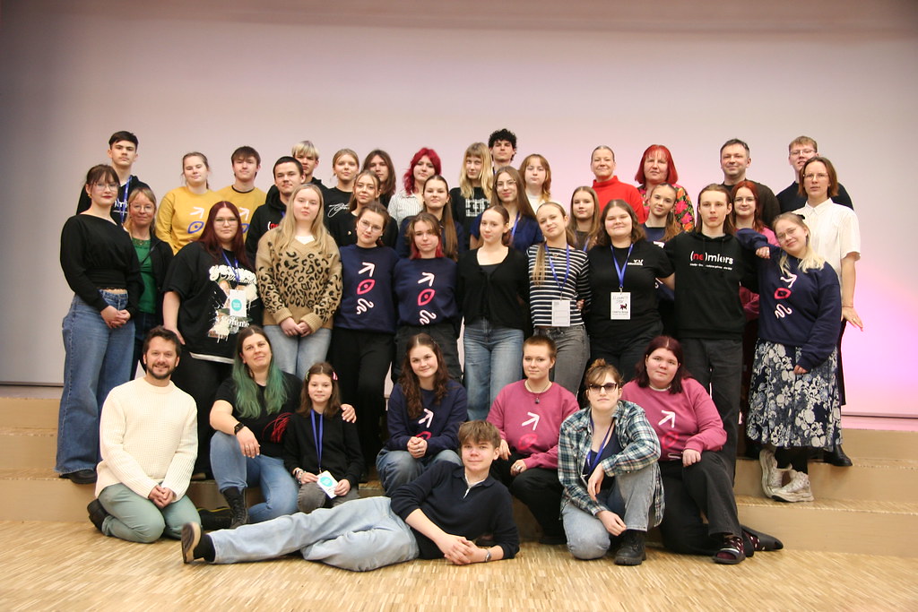 Liela, daudzveidīga aptuveni 40 jauniešu un dažu pieaugušo grupa, kas stāv un sēž vairākās rindās, smaidot kamerā. Daudzi valkā kreklus ar logotipiem un uzrakstiem. Vairāki cilvēki valkā tumši zilus vai rozā džemperus ar baltu vai rozā bultu, kas norāda pa labi, un stilizētu acu simbolu zem tās. Daži valkā melnus kreklus ar baltu uzrakstu "(ne)miers". Uz vienas kakla kartes redzams uzraksts "KLAPERIS" un suņa siluets. Fons ir gaiši pelēks ar rozā nokrāsu labajā pusē.