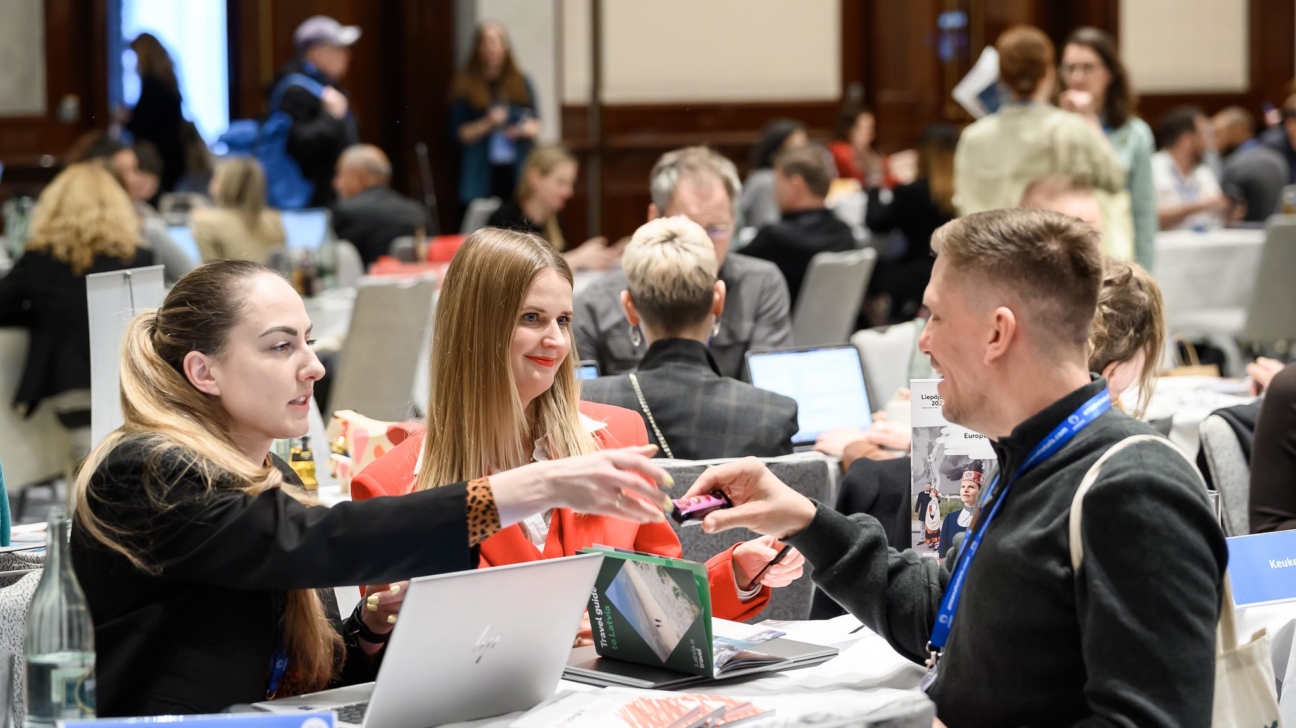 Divas sievietes un vīrietis sēž pie galda biznesa pasākumā. Sieviete sarkanā žaketē ar smaidu pasniedz vīrietim mazu, krāsainu priekšmetu. Vīrietis ir pasmaidījis. Uz galda blakus klēpjdatoram ir zila zīme ar tekstu "Latvia Travel" un brošūra ar uzrakstu "Travel guide" un "en Latvia". Fonā sēž citi pasākuma dalībnieki.