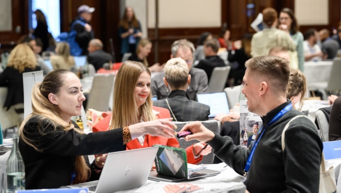 Sieviete oranžā žaketē smaidot pasniedz nelielu violetu priekšmetu smaidīgam vīrietim pie galda konferencē. Pie galda sēž arī cita sieviete, un uz tā redzami klēpjdatori, kā arī brošūra ar nosaukumu 