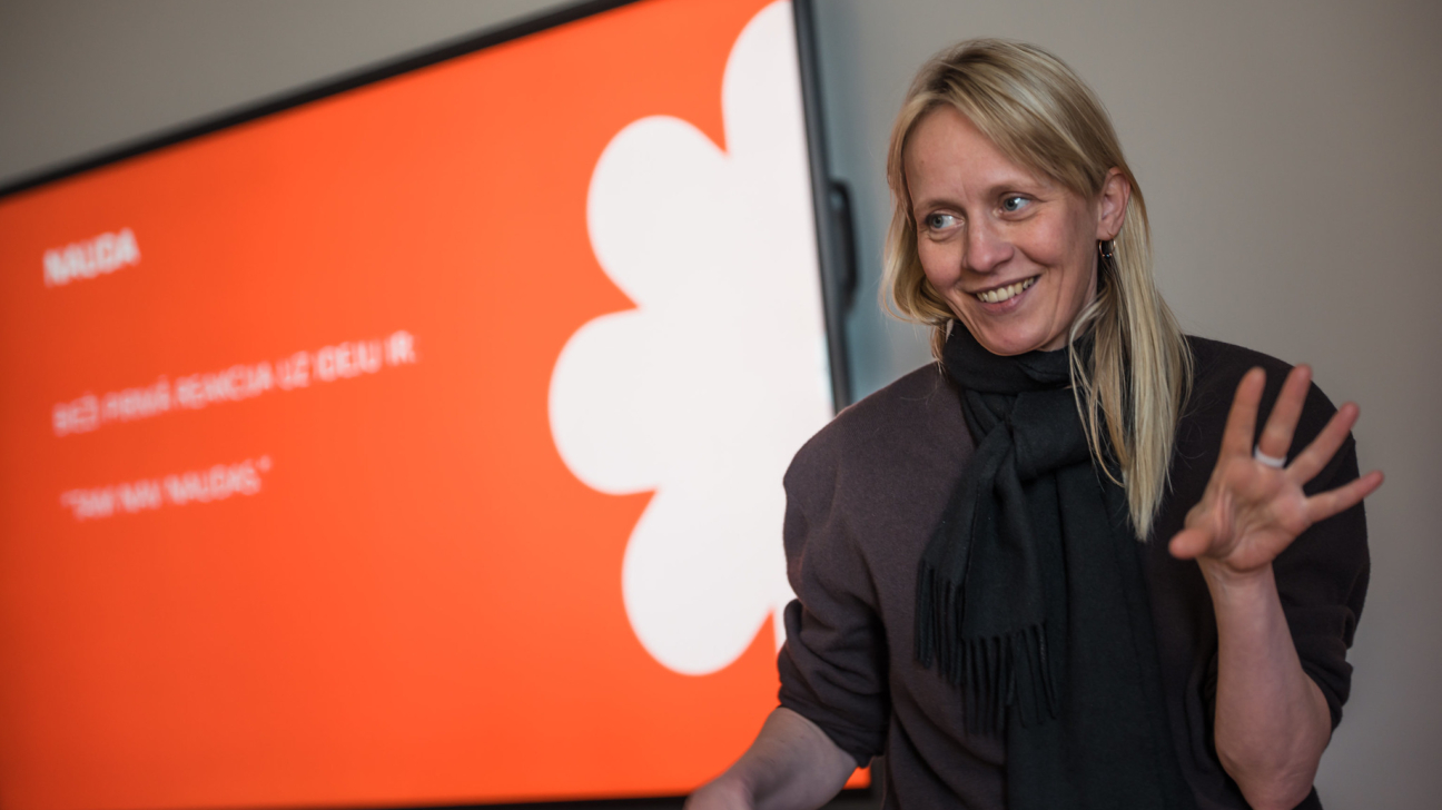 Sieviete ar šalli smaida un žestikulē ar paceltu labo roku. Aiz viņas oranžā ekrānā redzams balts stilizēta zieda attēls