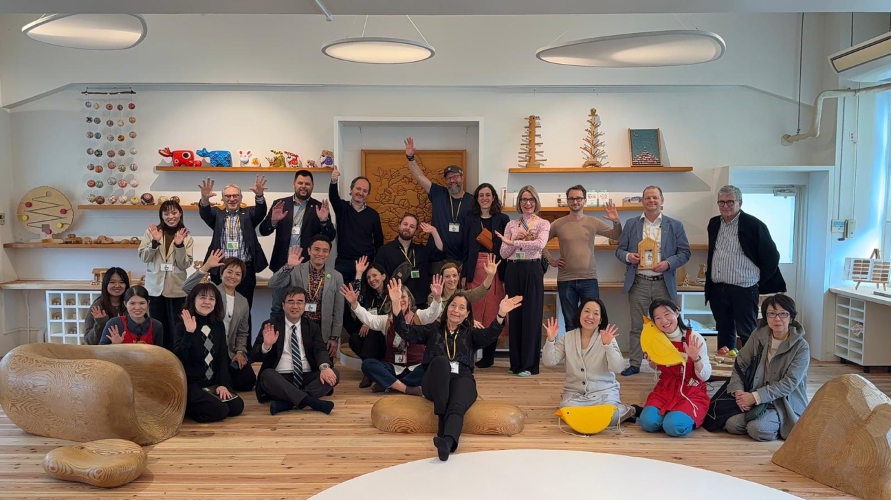 Liela, daudzveidīga cilvēku grupa pozē fotogrāfijai gaišā telpā ar koka grīdu un plauktiem, kas pildīti ar koka rotaļlietām un amatniecības izstrādājumiem. Lielākā daļa cilvēku smaida un vicina rokas; daži sēž uz liela izmēra koka skulptūrām un dzelteniem putnu formas sēdekļiem priekšplānā.