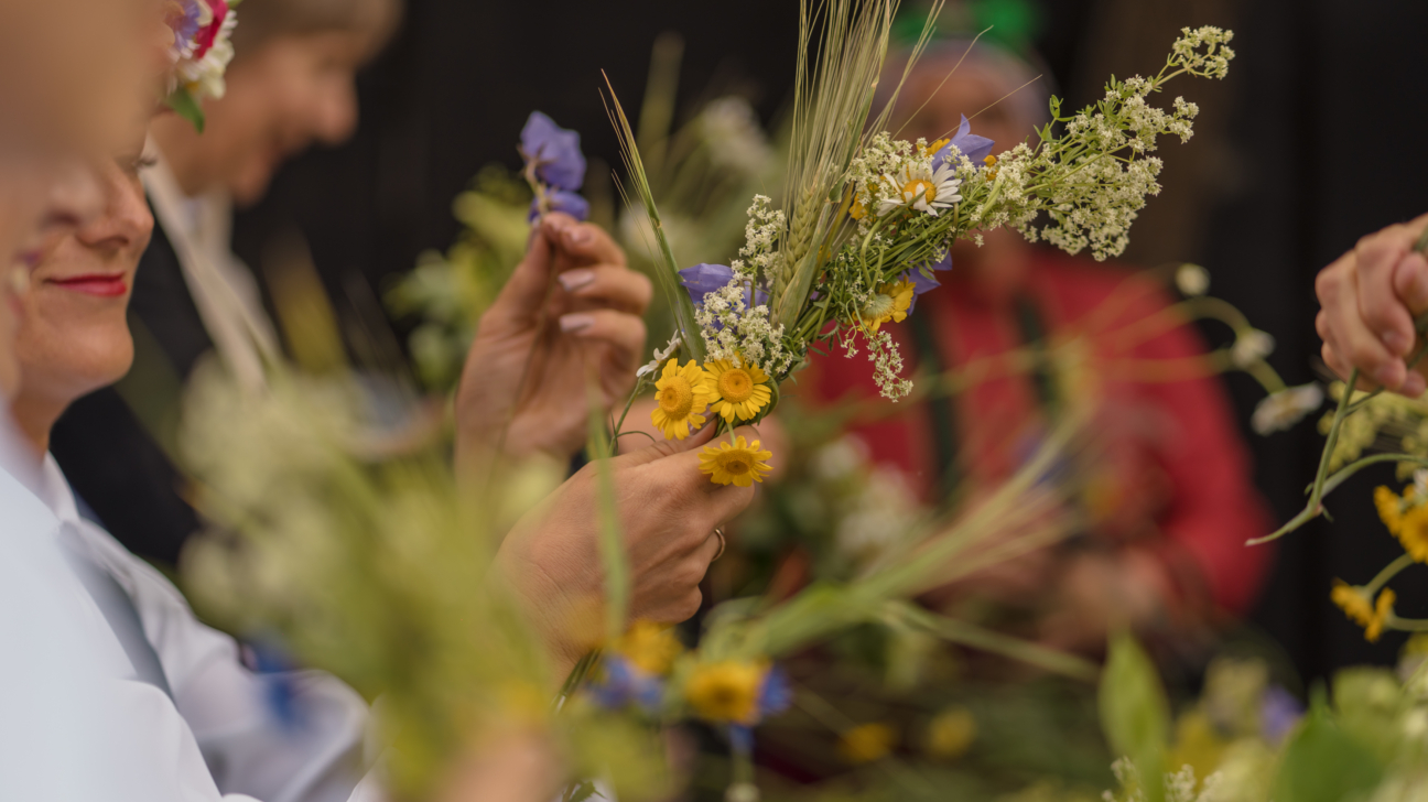Tuvplānā redzamas rokas, kas pin pušķi no rudzu vārpām, margrietiņām, sīkām baltām un zilām lauku puķēm. Fonā izplūduši citi cilvēki veido līdzīgus ziedu un graudaugu salikumus.