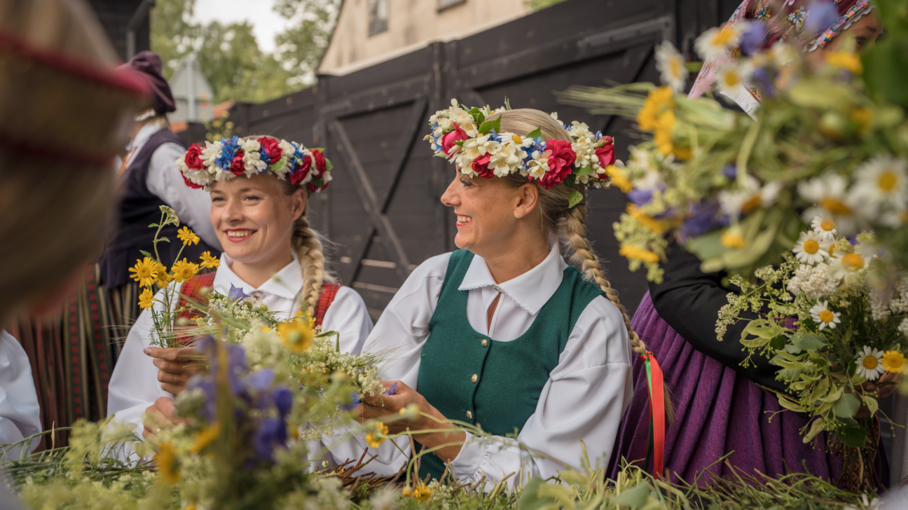 Divas smaidīgas sievietes latviešu tautastērpos ar ziedu vainagiem galvās pin vai kārto ziedus. Viņām ir pītas blondas bizes, un apkārt redzami dažādi pļavas ziedi.