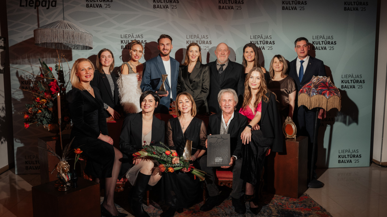 Cilvēku grupa, dažāda vecuma vīrieši un sievietes svinīgos tērpos, pozē kopbildei Liepājas Kultūras balva '25 pasākumā. Daži tur zelta statuetes, puķu pušķi un balvu plāksni. Fonā redzams uzraksts "LIEPĀJAS KULTŪRAS BALVA '25".