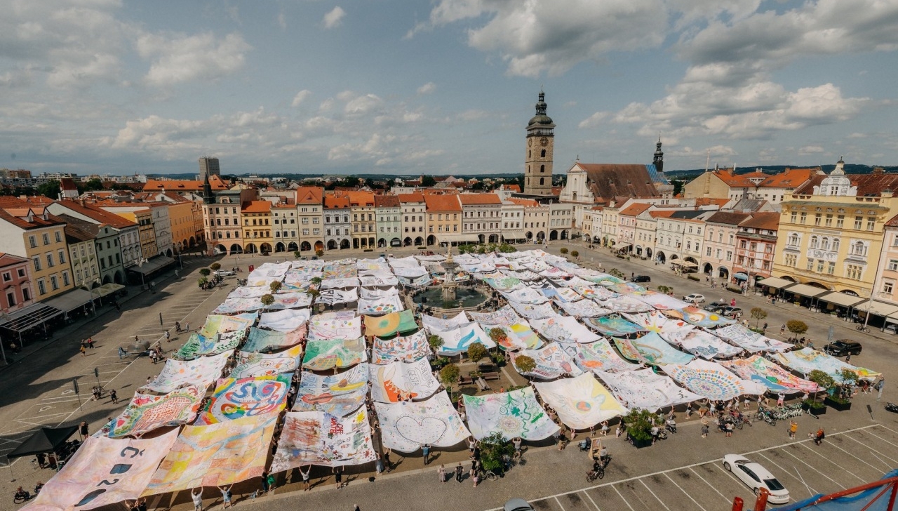 Augšupējs skats uz pilsētas laukumu, kur simtiem krāsainu, apgleznotu auduma gabalu ir izklāti un to malas tur rokās cilvēki, veidojot viļņainu instalāciju. Laukuma centrā ir strūklaka. Fonā redzamas vēsturiskas ēkas ar oranžiem jumtiem un augsts pulksteņa tornis zem zilām, daļēji mākoņainām debesīm.