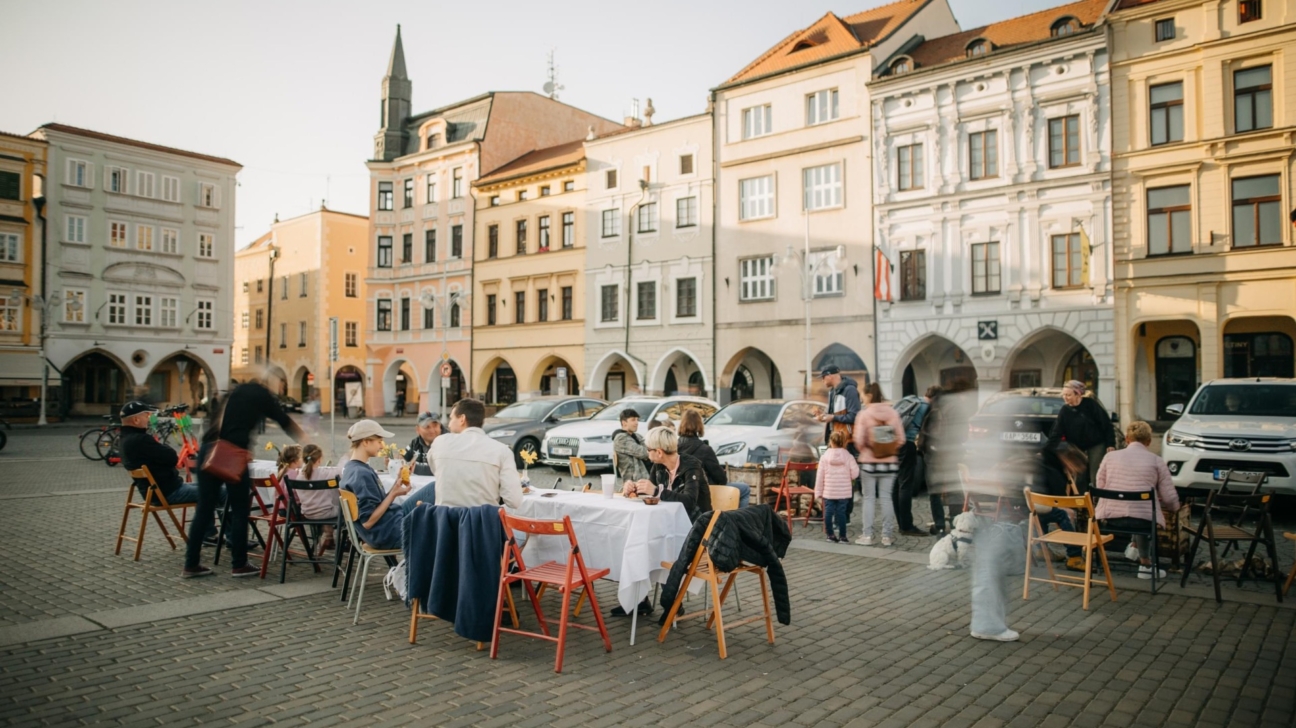 Dzīvīga aina bruģētā pilsētas laukumā, kur vairākas cilvēku grupas ietur maltīti ārā pie galdiem ar baltiem galdautiem. Fonā rindojas vēsturiskas daudzstāvu ēkas ar arkveida pirmajiem stāviem un novietotas automašīnas. Daži cilvēki pārvietojas, radot kustības izplūšanas efektu.