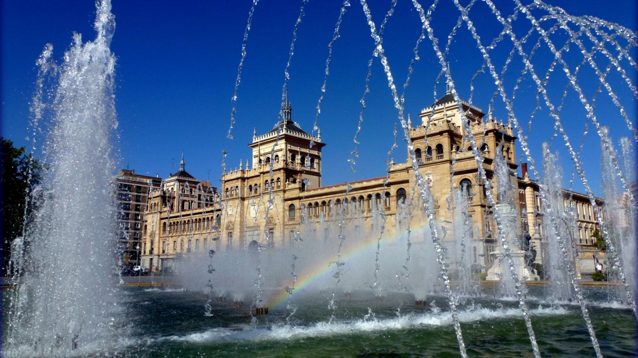 Liela strūklaka ar ūdens šļakatām, kurās redzama maza varavīksne, un aiz tās paceļas majestātiska, gaišas krāsas ēka ar torņiem zem zilām debesīm.