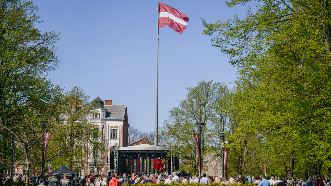 Pūlis pulcējies parkā pie skatuves ar paceltu Latvijas karogu centrā. Aiz muguras redzama vēsturiska ēka un zaļojoši koki saulainā dienā.