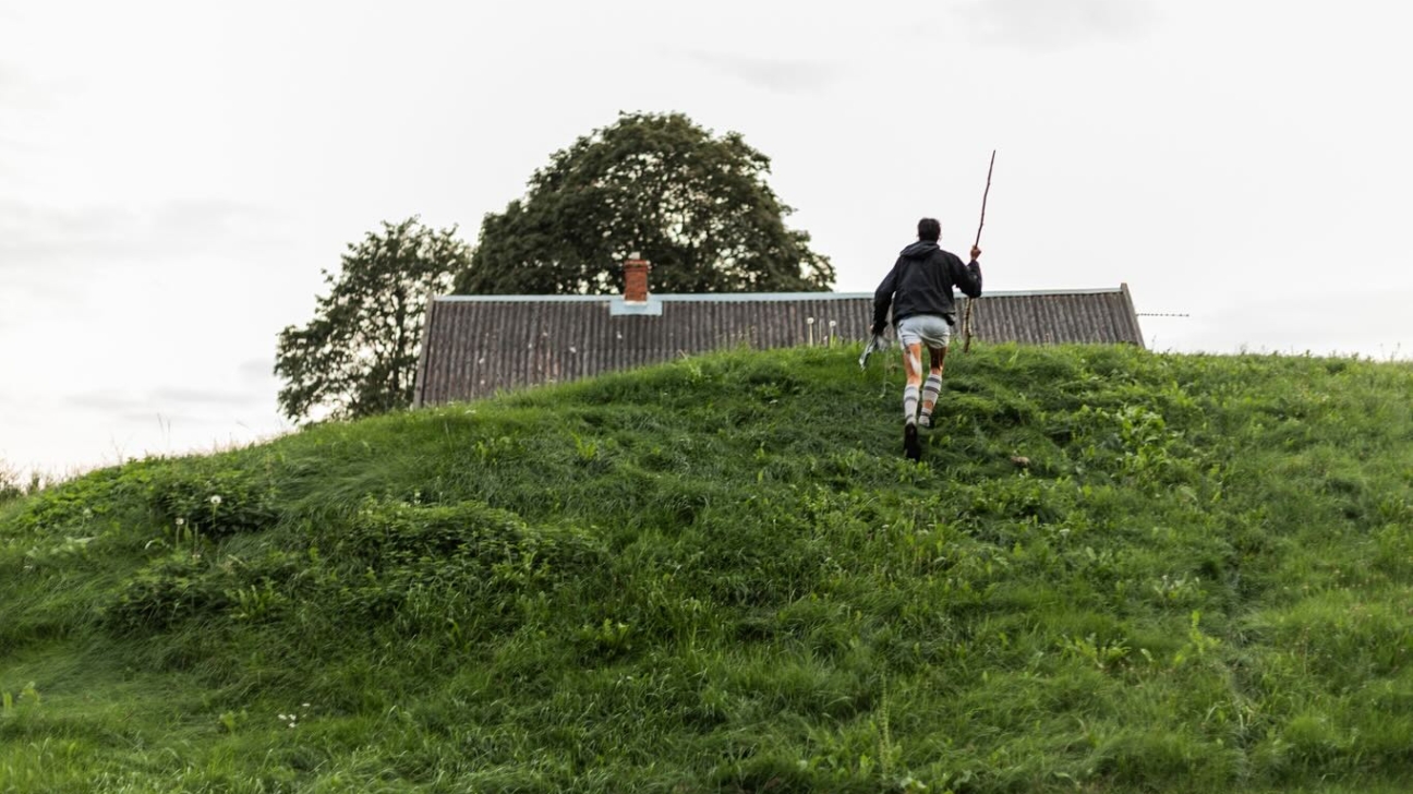 Cilvēks melnā jakā un gaišos šortos ar svītrainām kāju aptiniem kāpj augšup pa zālainu pakalnu, turot rokā koka zaru. Pakalna galā redzama māja ar pelēku jumtu un liels koks zem apmākušām debesīm.