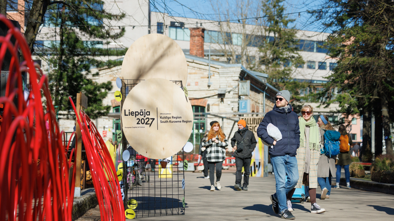 Saulaina diena pilsētā, kur cilvēki pastaigājas garām sarkanai mākslas instalācijai un stendiem ar informāciju. Uz diviem apļveida kokmateriālu plakātiem rakstīts: "Liepāja 2027 European Capital of Culture" un "Official partners Kuldīga Region South Kurzeme Region".