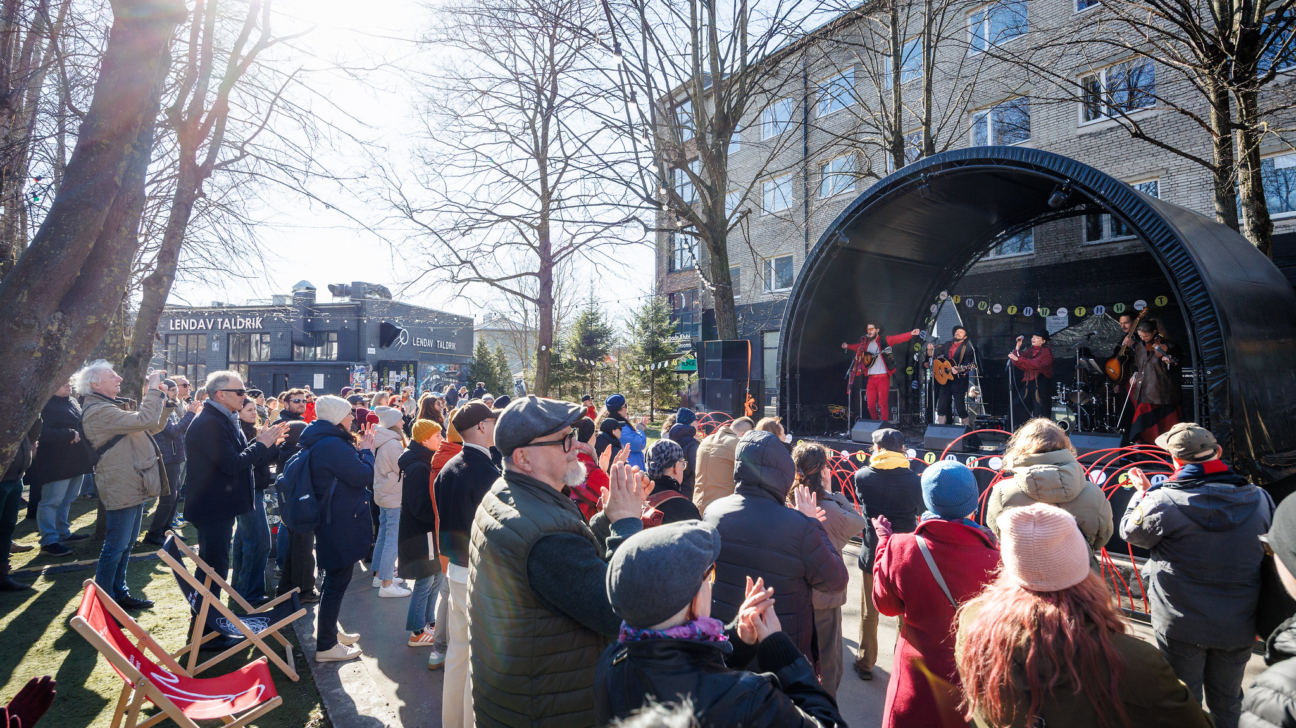 Cilvēku pūlis pulcējies ārā saulainā dienā, vērojot grupas uzstāšanos uz melnas, arkveida skatuves. Daži cilvēki aplaudē, citi fotografē ar telefoniem. Fonā redzama ēka ar uzrakstu "LENDAV TALDRiK".