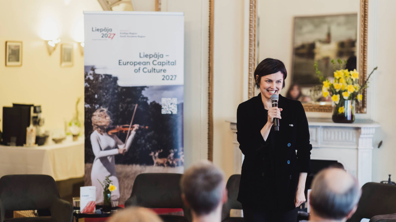 Smaidoša sieviete ar īsiem tumšiem matiem, ģērbusies melnā žaketē, tur mikrofonu un uzstājas. Aiz viņas ir reklāmas stends ar tekstu "Liepāja 2027", "Kurzeme Region", "South Kurzeme Region", "Liepāja – European Capital of Culture 2027" un attēlu ar sievieti, kas spēlē vijoli. Priekšplānā redzama auditorija.