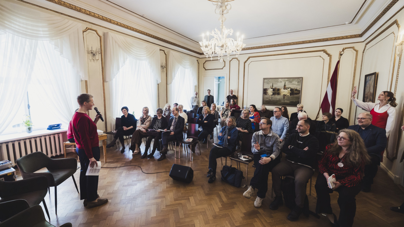 Persona sarkanā džemperī runā mikrofonā, stāvot priekšā aptuveni 30 cilvēku auditorijai, kas sēž krēslos greznā telpā ar augstiem griestiem un lieliem logiem. Aizmugurē labajā pusē redzams Latvijas karogs.