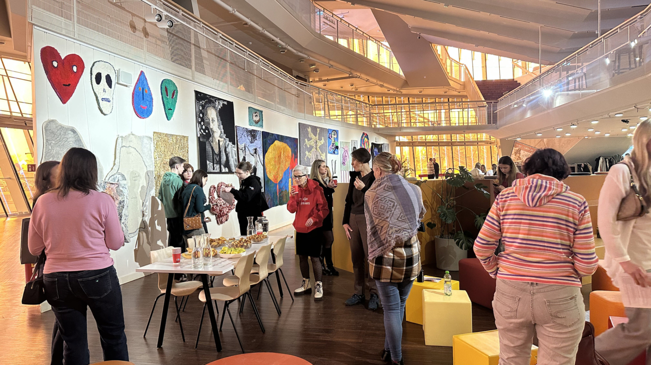 Grupa cilvēku atrodas plašā, modernā izstāžu telpā ar daudzlīmeņu arhitektūru. Uz baltas sienas izkārtoti dažādi mākslas darbi, tostarp tekstila sirdis, galvaskausi, sejas un abstrakcijas, kā arī melnbalts portrets. Cilvēki sarunājas un apskata ekspozīciju, daži stāv pie galda ar uzkodām. Uz grīdas redzams oranžs aplis ar uzrakstu "Piano Klusu".