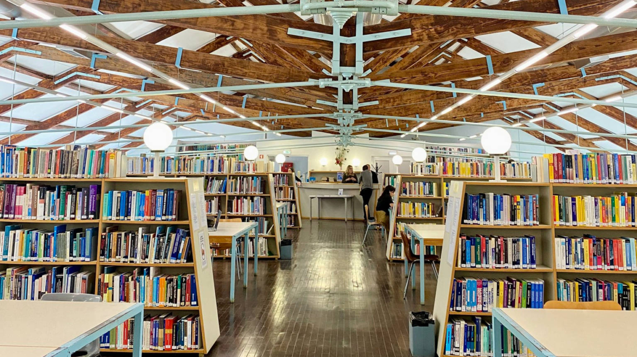 Plašs bibliotēkas interjers ar garām koka grāmatu plauktu rindām, kas piepildītas ar krāsainām grāmatām. Grīda ir no tumša koka parketa. Griestos redzama iespaidīga koka siju konstrukcija ar baltiem paneļiem un sfēriskiem gaismekļiem. Daži cilvēki atrodas pie apkalpošanas letes telpas galā, citi sēž pie galdiem.