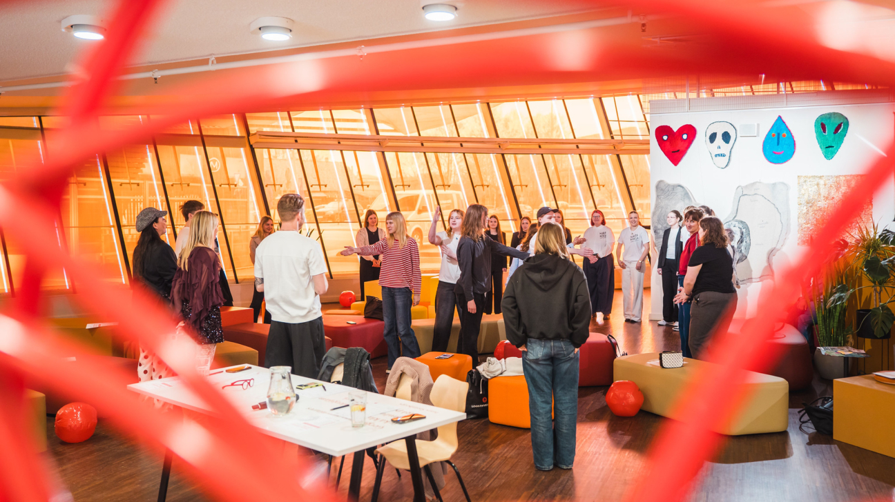 Caur sarkanu, abstraktu struktūru redzama moderna, gaiša telpa, kurā apmēram piecpadsmit jauni cilvēki, galvenokārt sievietes, stāv un iesaistās diskusijā, daži žestikulē. Telpai ir lieli oranži logi un uz sienas ir sarkana sirds, balts galvaskauss, zils piliena un zaļa citplanētieša galva. Priekšplānā redzams galds ar glāzēm un papīriem.