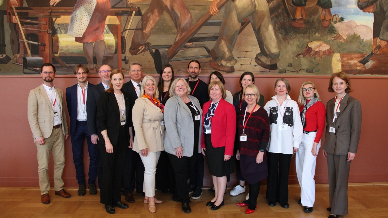 Grupa, kurā ir aptuveni 17 profesionāli ģērbti pieaugušie – vīrieši un sievietes –, smaida un pozē fotogrāfijai telpās. Viņi stāv uz koka grīdas, priekšā lielai, krāsainai sienas gleznai, kas attēlo cilvēkus darbībā, un sarkanbrūnai sienai.