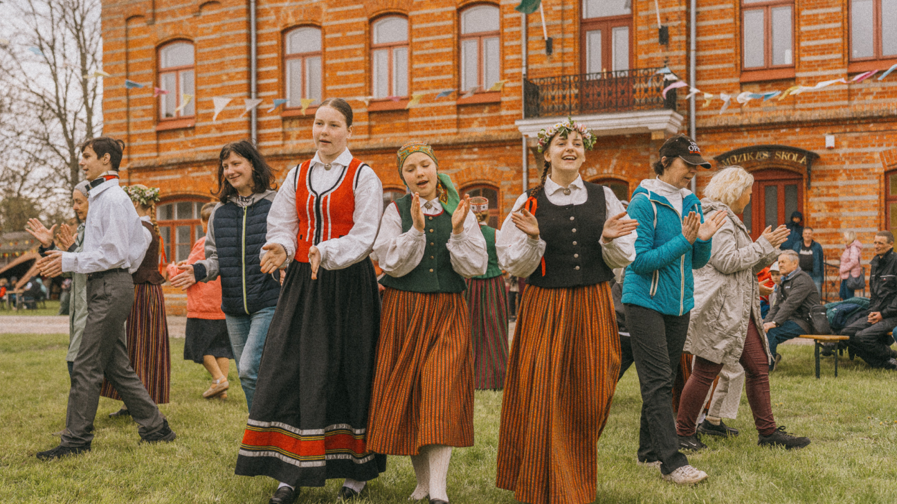 Grupa cilvēku, tostarp vairākas jaunas sievietes latviešu tautas tērpos un ziedu vainagos, dejo un aplaudē uz zāles laukuma. Fonā redzama sarkana ķieģeļu ēka ar uzrakstu "MŪZIKAS SKOLA" un Latvijas karogu.