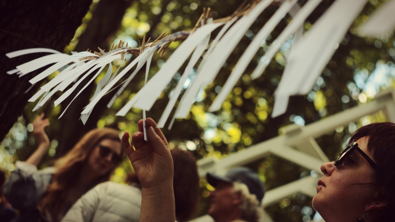 Cilvēka roka sniedzas pēc baltas papīra strēmeles, kura kopā ar citām līdzīgām strēmelēm ar veļas knaģiem piestiprināta pie koka zara. Priekšplānā sieviete skatās uz augšu, fonā izplūduši koki un citi cilvēki.