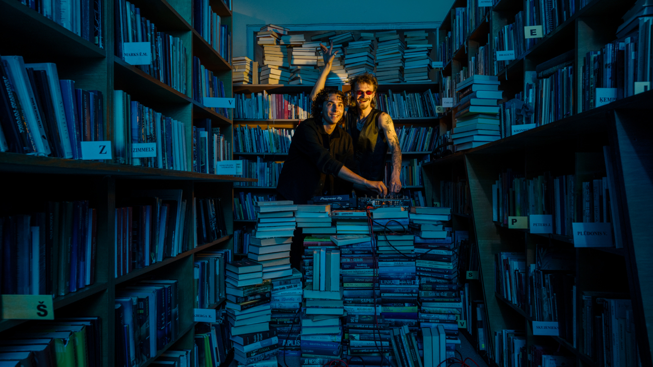 Divi smaidīgi vīrieši stāv starp grāmatu plauktiem bibliotēkā vai grāmatnīcā, kas ir apgaismota zilā gaismā. Viens vīrietis ar sarkanām saulesbrillēm un tetovētu roku paceļ vienu roku, otrs vīrietis apkalpo "Pioneer DJ" pulti, kas atrodas uz grāmatu kaudzes. Plauktos ir tūkstošiem grāmatu, un uz dažiem plauktiem redzamas uzlīmes, piemēram, "MARKS E.M.", "ZIMMELS", "ELSBERGS S.", "KEMPS J.", "PETERS J.", "PLUDONIS V.". Uz grīdas ir sarkans vads.