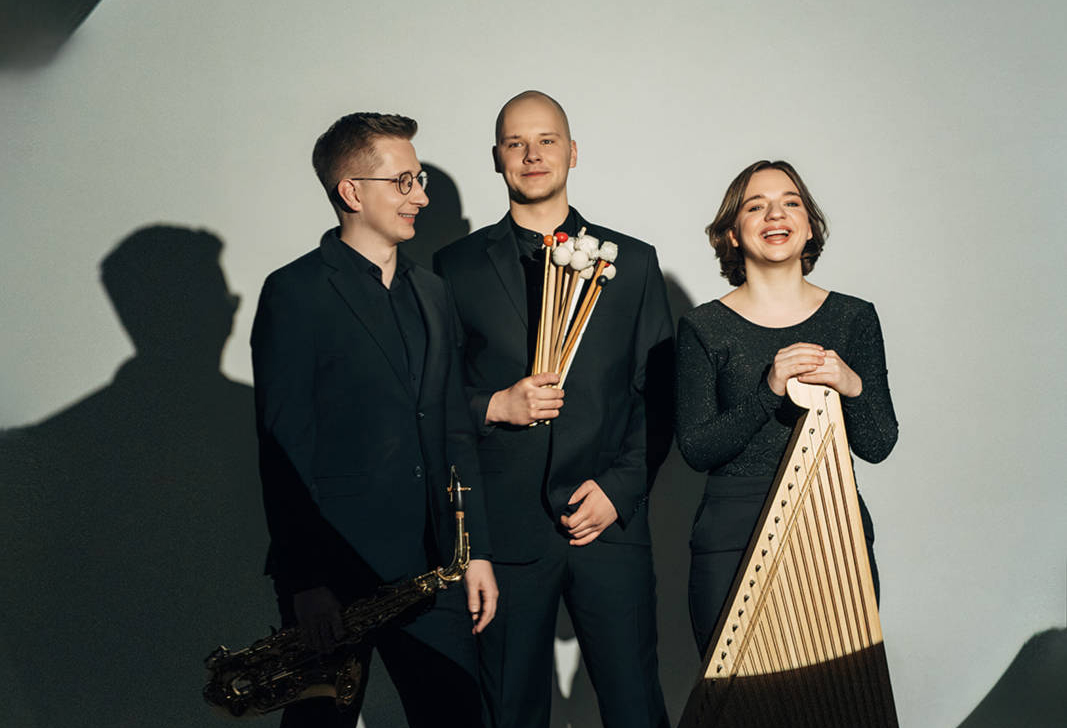 Trīs smaidīgi mūziķi, ģērbušies tumšā apģērbā, stāv pie gaišas sienas ar garām ēnām. Vīrietis pa kreisi tur saksofonu, vīrietis centrā tur perkusiju vālītes, un sieviete pa labi tur kokli.