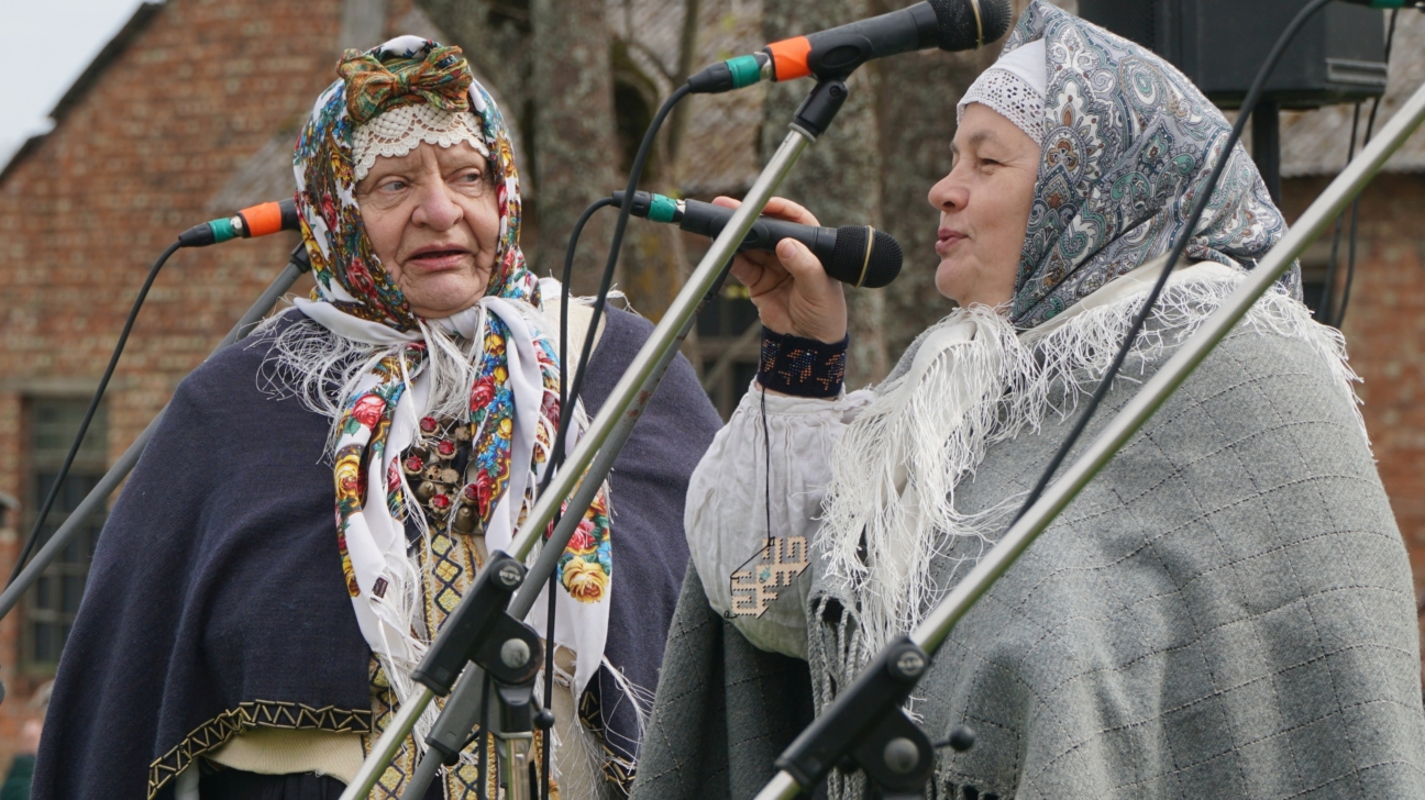 Divas sievietes tradicionālos tērpos ar galvas lakatiem un šallēm dzied mikrofonos brīvdabas pasākumā.