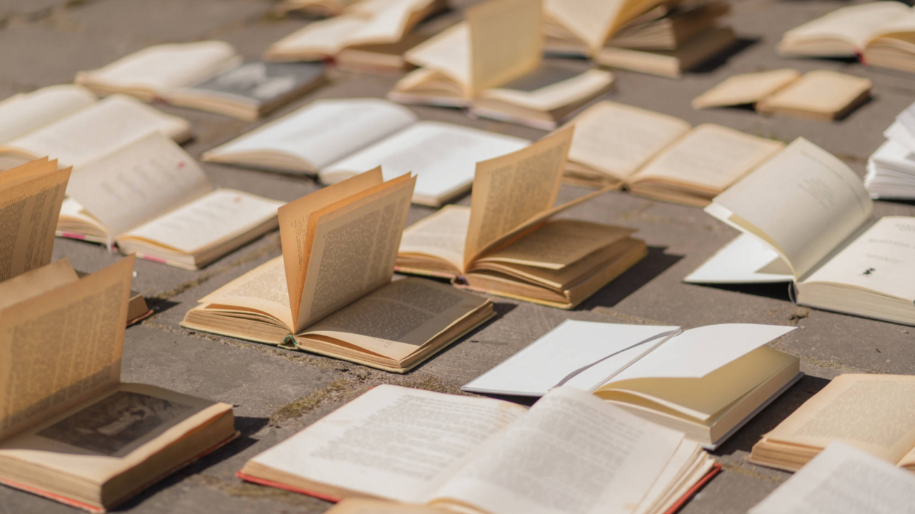 Daudzas vecas, atvērtas grāmatas ar dzeltenīgām lapām izkaisītas uz saulainas bruģētas virsmas, metot ēnas.