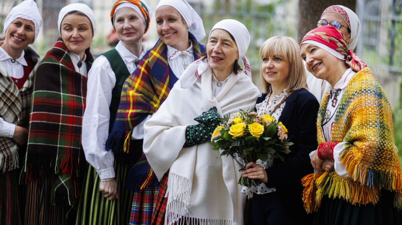 Deviņas sievietes, tērpušās krāsainos latviešu tautastērpos ar rakstainām villainēm un galvas lakatiem, smaida un pozē ārā. Viena no sievietēm tur dzeltenu rožu pušķi.