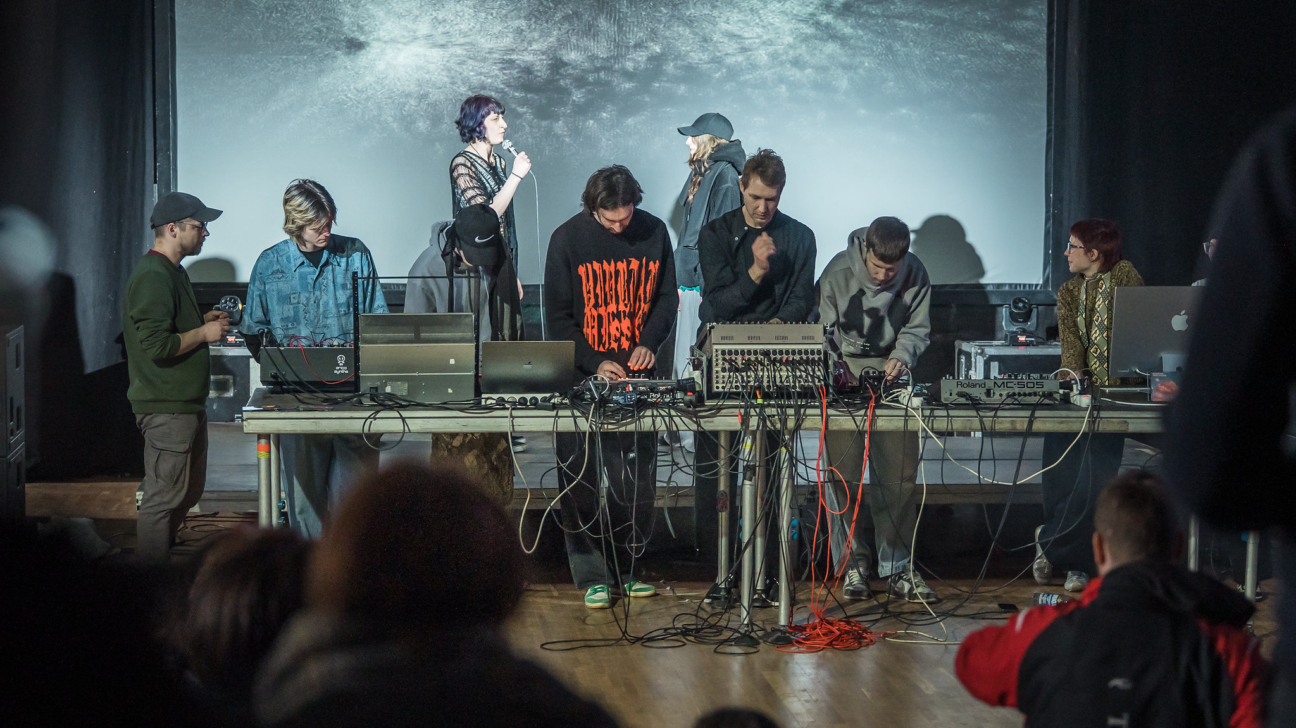 Grupa cilvēku uz skatuves veic performanci ar elektronisko mūzikas aprīkojumu. Sieviete ar violetiem matiem runā mikrofonā. Pie gara galda vairāki cilvēki pielāgo mikserus, datorus un citas ierīces, tostarp "Roland MC-505". Uz viena melnā džempera ir uzraksts "ANCIENT GRAVES". Aizmugurē uz liela ekrāna redzama abstrakta vizualizācija, priekšplānā – skatītāji.
