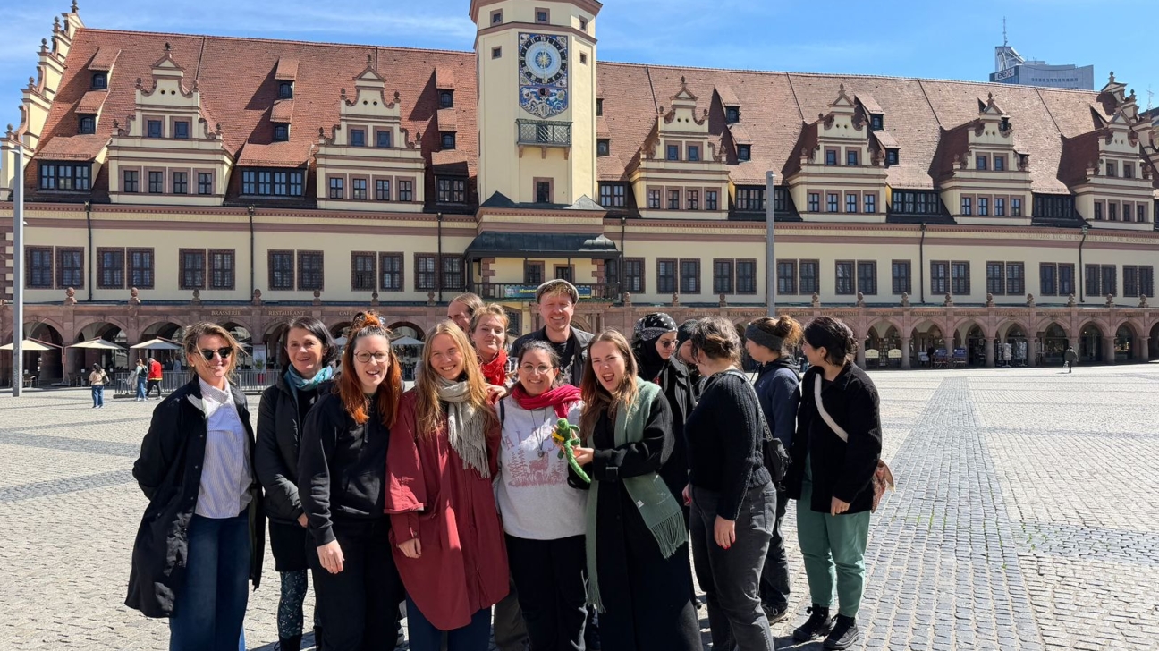 Apmēram 15 jauniešu grupa smaida, pozējot fotogrāfijai saulainā dienā. Viņi stāv uz bruģēta laukuma, priekšā plašai vēsturiskai ēkai ar sarkanu dakstiņu jumtu un augstu pulksteņa torni ar zaļu kupolu. Uz ēkas fasādes virs arkām redzami uzraksti "BRAUERIE", "RESTAURANT", "CAFE".