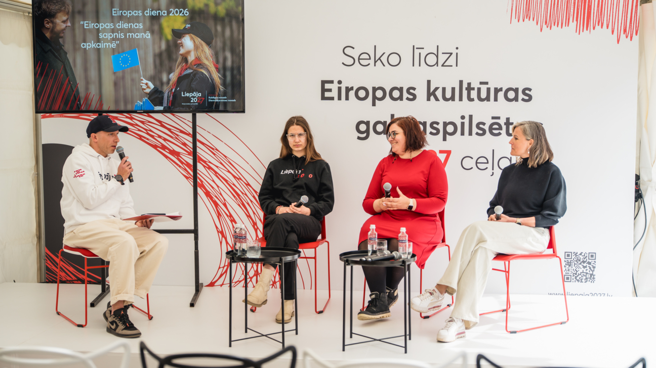 Četri cilvēki – vīrietis un trīs sievietes – sēž uz skatuves sarkanos krēslos, piedaloties diskusijā. Vīrietis runā mikrofonā. Fona ekrānā redzams uzraksts “Eiropas diena 2026” un “Eiropas dienas sapnis manā apkaimē”, zem tā Liepāja 2027 logo un attēls ar cilvēkiem, kas tur ES karogu. Aizmugurējā sienā lasāms “Seko līdzi Eiropas kultūras galvaspilsēta 2027 ceļā”.