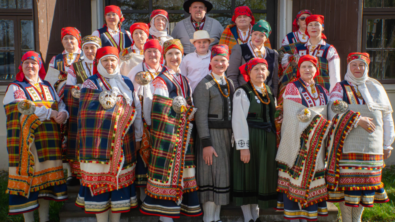 Liela grupa cilvēku, tostarp sievietes, vīrieši un zēns, tērpušies krāsainos tautas tērpos ar izšuvumiem, galvas lakatiem un saktām, stāv ārā pie koka ēkas.