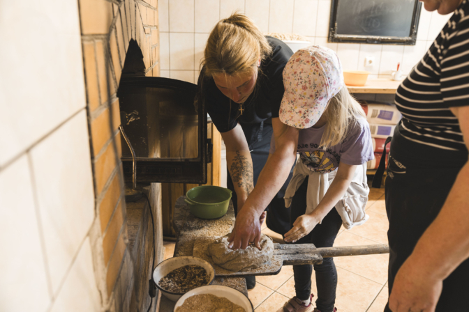 Sieviete un meitene pie ķieģeļu maizes krāsns mīca mīklu uz koka lāpstas. Blakus ir bļodas ar graudaugiem. Meitenei ir ziedu raksta cepure, un uz t-krekla redzams uzraksts "MAKE LOVE NOT WAR". Daļēji redzama vēl viena persona.