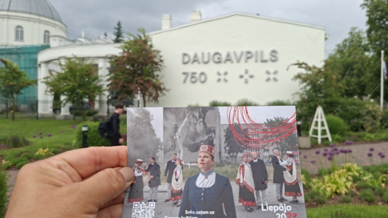 Roka tur pastkarti ar uzrakstiem "Seko ceļam uz Eiropas kultūras galvaspilsētu 2027", "Liepāja 2027", "Eiropas kultūras galvaspilsēta" un QR kodu, fonā redzama balta ēka ar uzrakstu "DAUGAVPILS 750" un kupolveida ēka.
