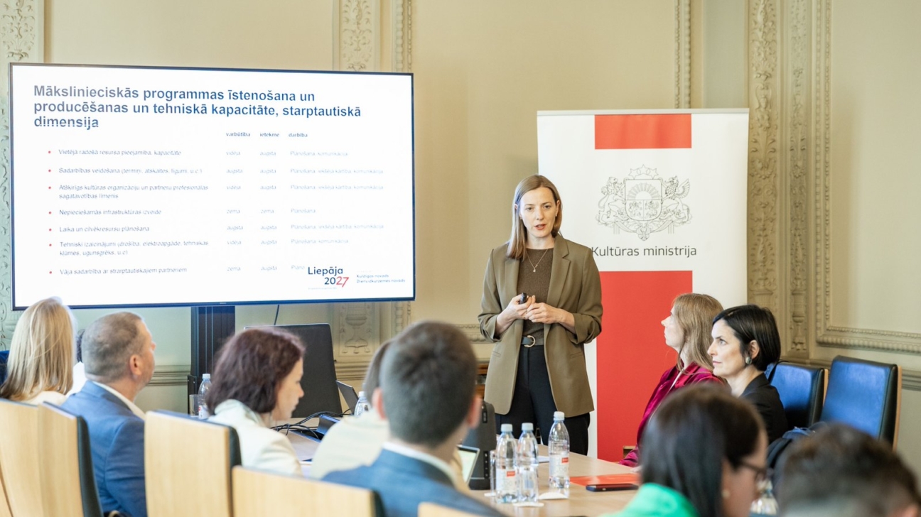 Sieviete prezentē auditorijai telpā ar lielu ekrānu, kurā redzams virsraksts "Mākslinieciskās programmas īstenošana un producēšanas un tehniskā kapacitāte, starptautiskā dimensija" un "Liepāja 2027". Aiz viņas ir Kultūras ministrijas reklāmkarogs, un vairāki cilvēki sēž pie galda, klausoties.