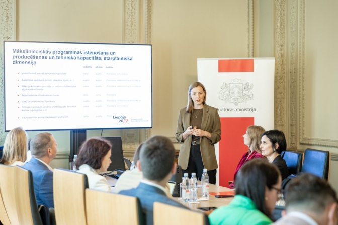 Sieviete prezentē auditorijai telpā ar lielu ekrānu, kurā redzams virsraksts "Mākslinieciskās programmas īstenošana un producēšanas un tehniskā kapacitāte, starptautiskā dimensija" un "Liepāja 2027". Aiz viņas ir Kultūras ministrijas reklāmkarogs, un vairāki cilvēki sēž pie galda, klausoties.
