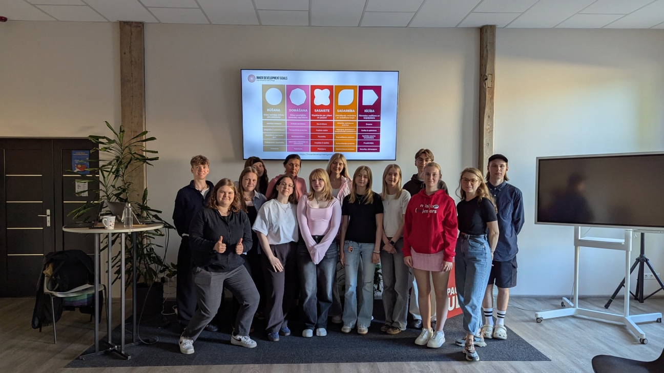 Grupa ar aptuveni 15 jauniem cilvēkiem stāv telpā. Fonā liels ekrāns ar virsrakstu "INNER DEVELOPMENT GOALS" un piecām krāsainām sadaļām – "BŪŠANA", "DOMĀŠANA", "SASAISTE", "SADARBĪBA", "RĪCĪBA" – katra ar apakšpunktiem. Kreisajā pusē viena sieviete rāda divus īkšķus uz augšu.