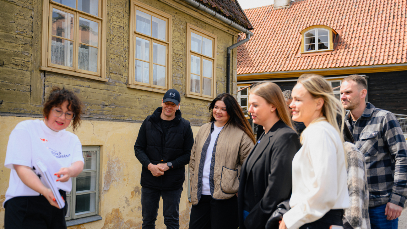 Seši cilvēki stāv ārā pie vecas koka ēkas ar dzeltensarkanu sienu un dakstiņu jumtu. Sieviete kreisajā pusē, ģērbusies baltā kreklā ar uzrakstu "Savā pilsētā", smaidot rāda kaut ko mapē pārējiem pieciem cilvēkiem, kas viņā uzmanīgi klausās.