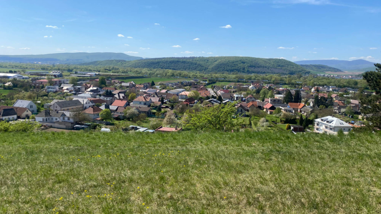 Skatījums no zāļaina pakalna uz mazpilsētu ar daudzām mājām un kokiem. Fonā ir zaļi, mežaini kalni zem skaidrām zilām debesīm.