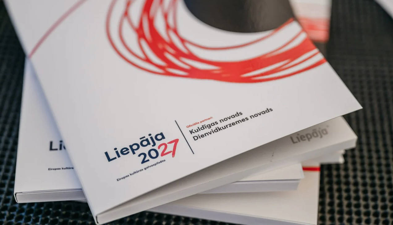 Baltu brošūru kaudze. Uz augšējās brošūras vāka redzams teksts "Liepāja 2027 Eiropas kultūras galvaspilsēta" un "Oficiālie partneri: Kuldīgas novads Dienvidkurzemes novads", ko papildina sarkans, abstraktas formas dizains.