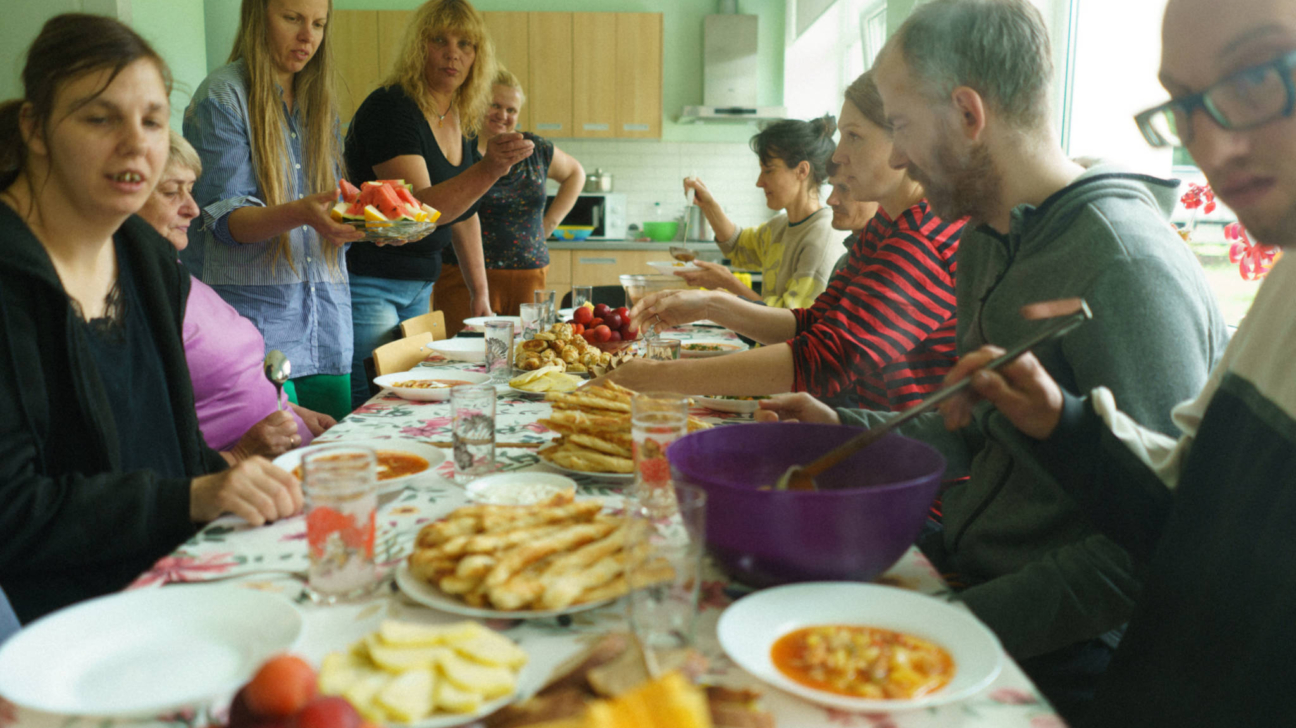 Cilvēku grupa pulcējusies ap galdu, kas klāts ar ziedainu galdautu un dažādiem ēdieniem, tostarp arbūza šķēlēm, siera standziņām un zupu. Vairāki cilvēki ēd un pasniedz maltīti, kamēr citi sarunājas.
