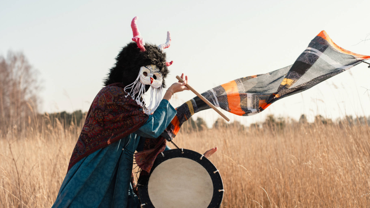Persona tradicionālā tērpā ar melnu kažokādas kapuci, rozā ragiem un baltu pūces masku stāv sausā zāles laukā. Viņa ir tērpusies zilā kleitā un sarkanā šallē, un tur lielu bungu un bungas vālīti, pie kuras piestiprināts rūtains, oranži melns audums, kas plīvo vējā.