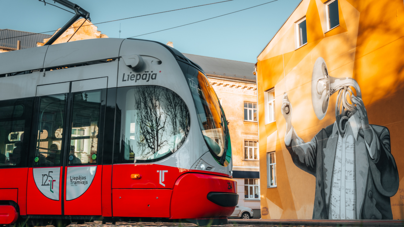Sarkans un balts tramvajs ar uzrakstu "Liepāja" priekšpusē un "125 Liepājas Tramvajs" sānos brauc garām oranžai ēkai. Uz ēkas sienas ir liels sienas gleznojums, kurā attēlota figūra ar tūbu galvas vietā un notīm rotātu kreklu, kas ar labo roku it kā diriģē.