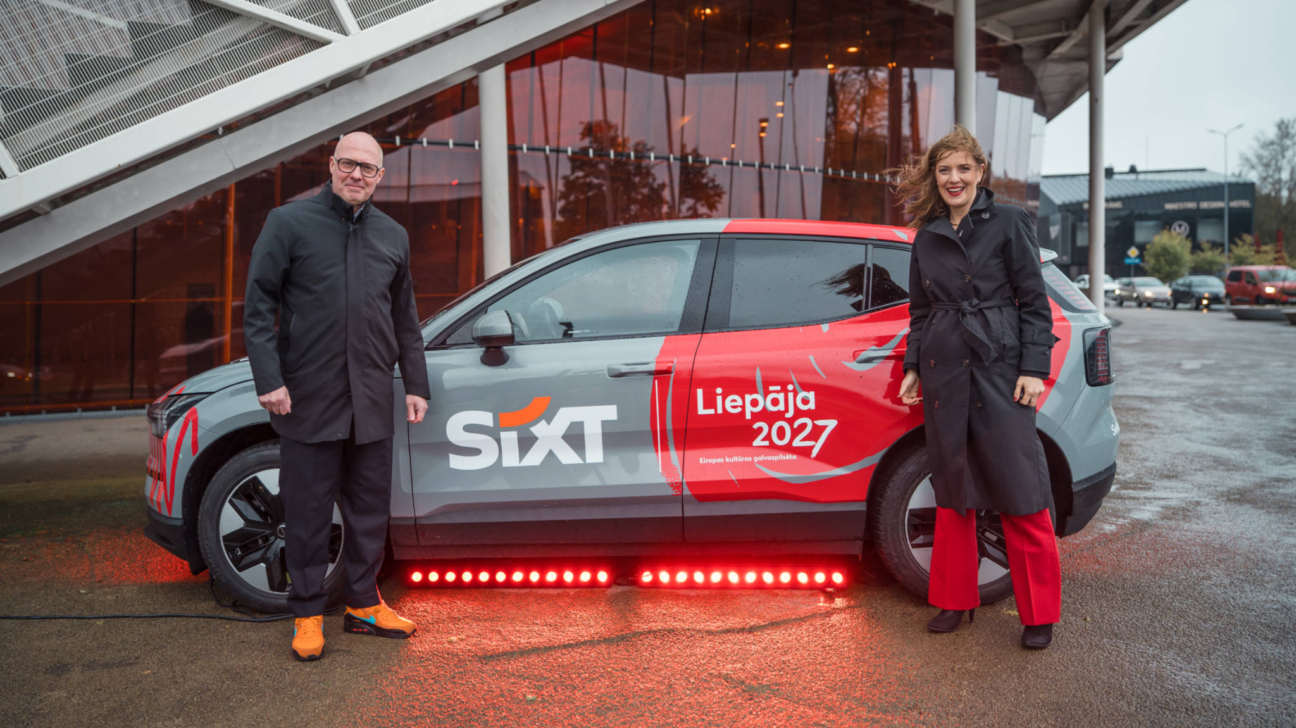 Vīrietis melnā mētelī un sieviete melnā mētelī un sarkanās biksēs stāv blakus pelēkai un sarkanai automašīnai ar uzrakstiem "SIXT" un "Liepāja 2027 Eiropas kultūras galvaspilsēta". Zem automašīnas ir sarkanas LED gaismas. Fonā redzama moderna ēka.