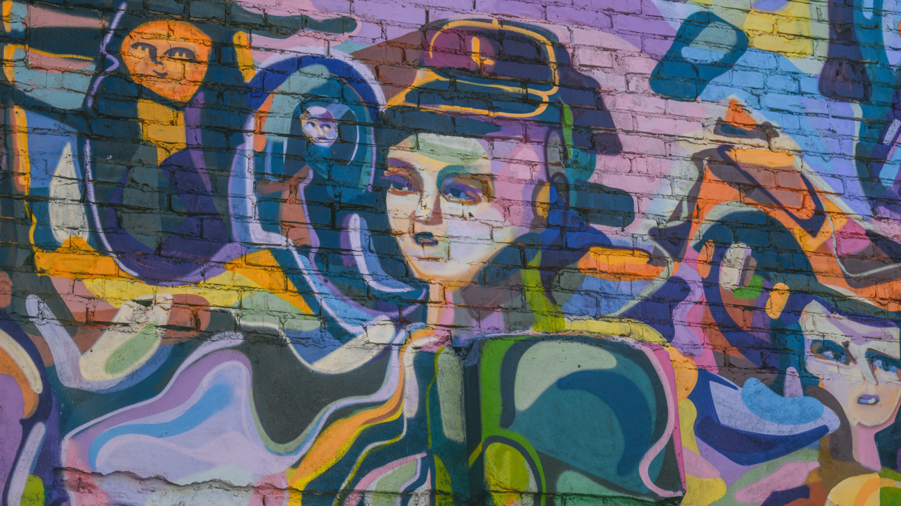 Krāsains grafiti uz ķieģeļu sienas, kurā centrā attēlota stilizēta sievietes seja cepurē, ko ieskauj abstraktas formas un citas daļējas figūras košos zilos, violetos, oranžos, dzeltenos un zaļos toņos.
