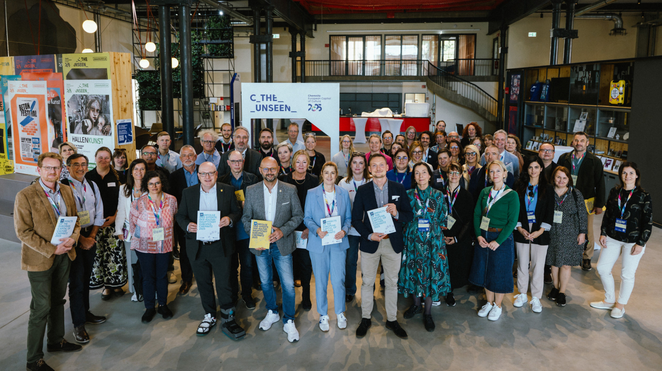 Liela pieaugušo grupa stāv smaidot industriāla stila telpā. Daži priekšplānā tur dokumentus ar uzrakstu "IMPACTS OF THE EUROPEAN CULTURAL CAPITAL CHEMNITZ 2025". Fonā redzams balts stends ar tekstu "C_THE_UNSEEN_ _ CHEMISTRY OF CULTURAL CAPITAL 2025".